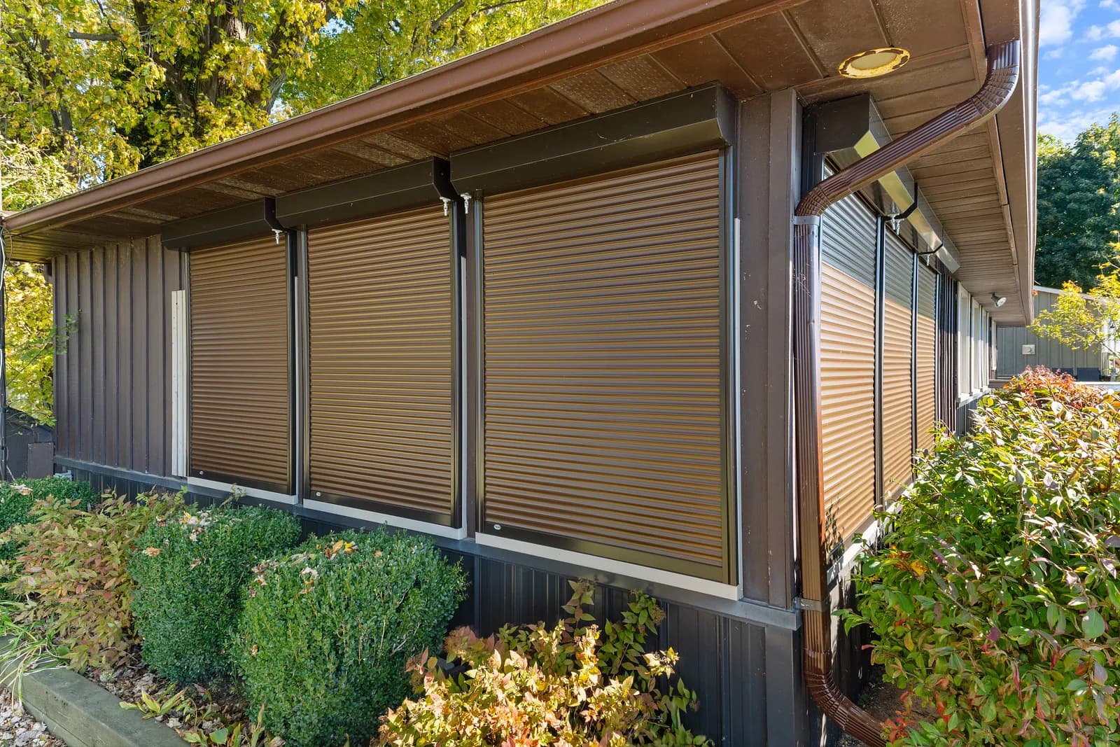 Three brown wood-grain aluminum roll shutters deployed across the side of a dark-clad home