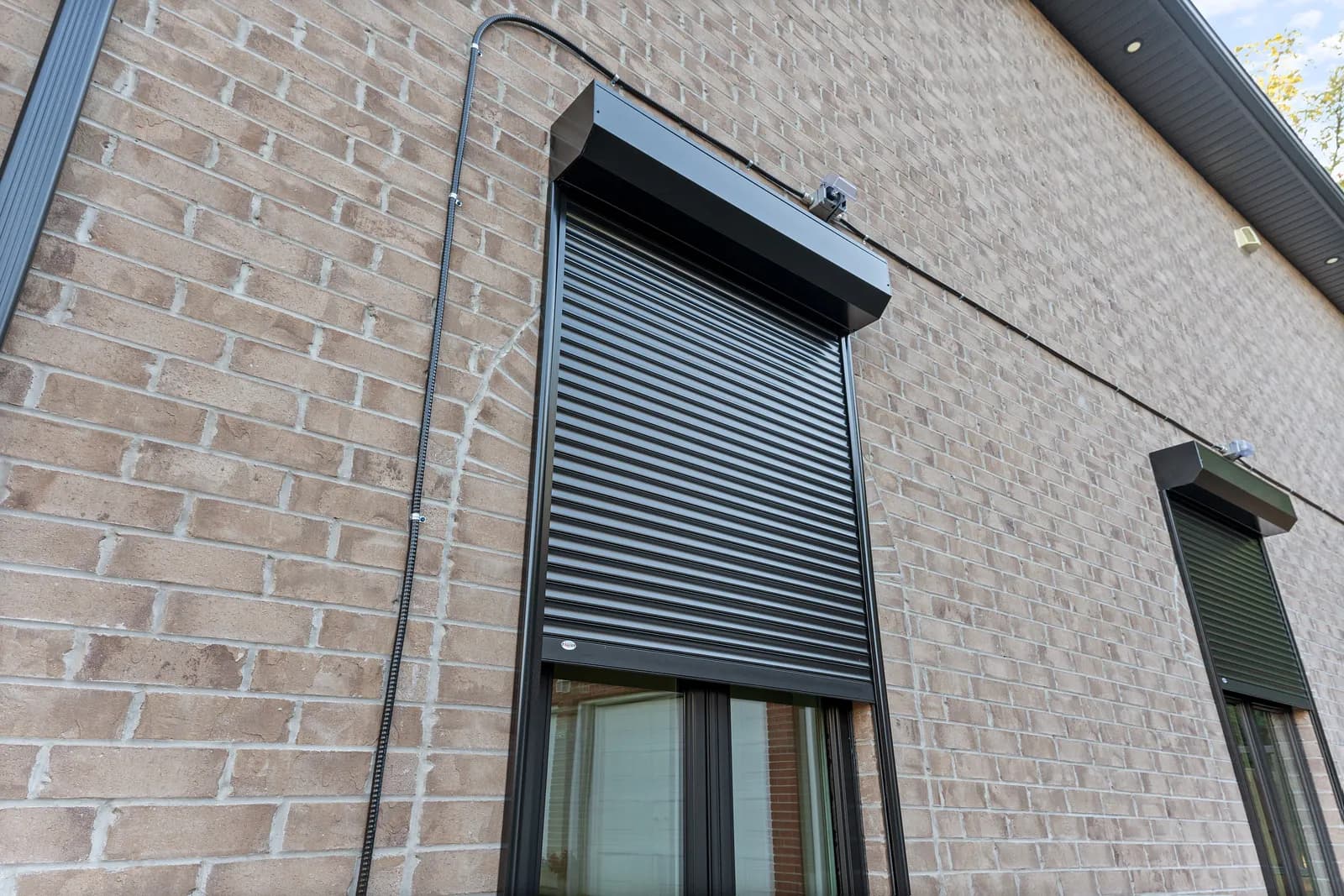 Tall black aluminum roll shutter deployed over a window on a brick wall beside a garden gate
